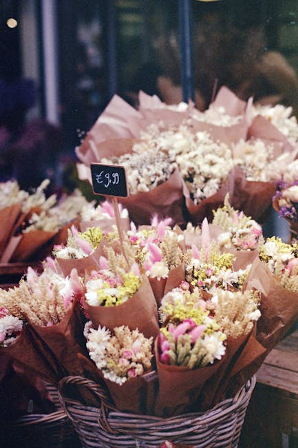 A basket filled with multiple small bouquets of fresh flowers, each wrapped in brown and pink paper. The arrangements feature pastel pink and cream blooms, including roses, carnations, and baby's brea