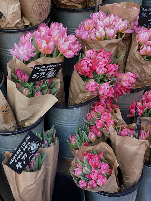 A display of various pink tulip bouquets arranged in brown paper wrapping, placed inside metallic buckets at a florist stand. The tulips exhibit shades ranging from soft pastel pink to vibrant magenta