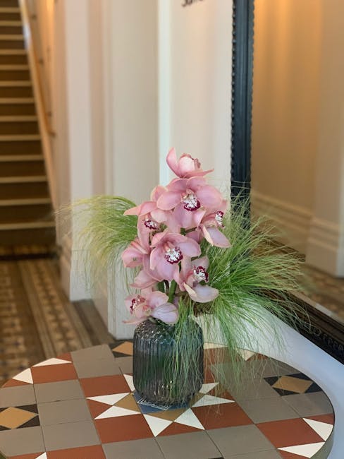 A tall, elegant floral arrangement featuring pink orchids with delicate petals and intricate white and purple markings in the center, set in a dark ribbed glass vase. The orchids are complemented by f
