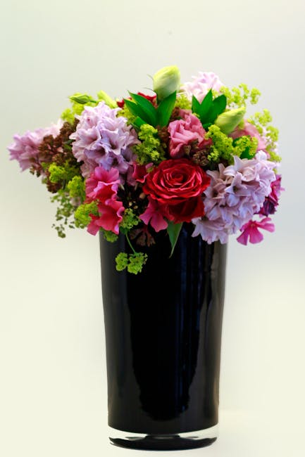 A tall, elegant black glass vase holding a lush floral bouquet featuring a variety of fresh flowers in soft pink, lavender, and bright red hues. The arrangement includes large roses, delicate hydrange