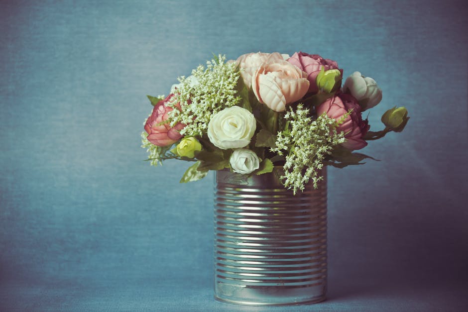 A floral arrangement featuring pink and white eustoma (lisianthus) and white astilbe flowers, artistically arranged in a cylindrical, ribbed metallic tin vase. The flowers are fresh, with delicate pet