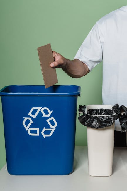 A person wearing a white shirt is disposing of floral foam into a blue recycling bin with a white recycling symbol, with a second container lined with a black trash bag nearby. The background features