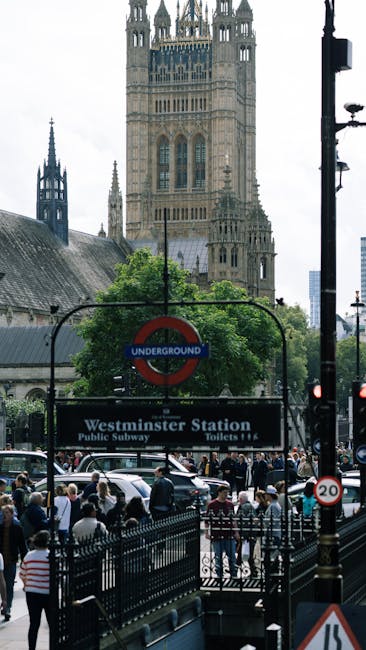 A busy scene outside Westminster Station in London, featuring a large black and white sign with the station name and subway information, set against a backdrop of historic architectural features of th