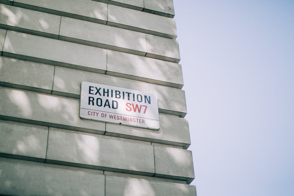 A close-up of a white building wall featuring a rectangular street sign with black and red text reading 'Exhibition Road SW7, City of Westminster.' The sign is mounted on a textured stone surface with