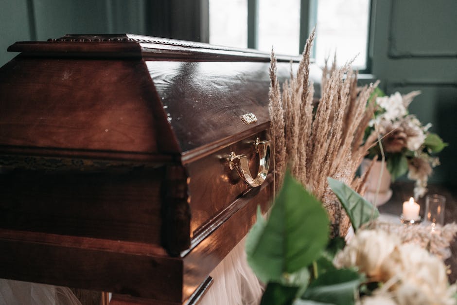 A polished wooden casket with a glossy finish and gold-colored handles is surrounded by floral arrangements and dried grasses. The flowers include white and blush pink blooms, arranged in vases and bo