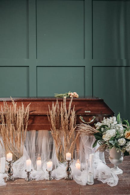 A floral arrangement featuring white and cream roses, peonies, and greenery placed in a white ceramic vase, positioned on the right side of a polished wooden coffin. Surrounding the coffin are tall, d