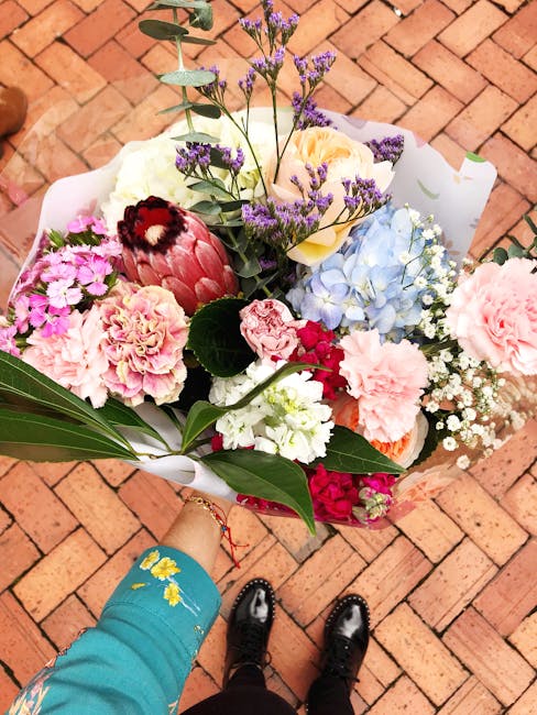 A vibrant bouquet of fresh flowers held by a person wearing a turquoise sleeve with yellow floral embroidery, positioned above a brick pavement. The arrangement includes pink carnations, white roses, 
