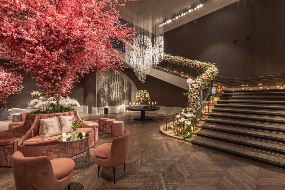 An elegant indoor floral display featuring a large, vibrant pink cherry blossom tree with numerous delicate blossoms. The arrangement includes smaller white and pink flowers in vases placed on a round