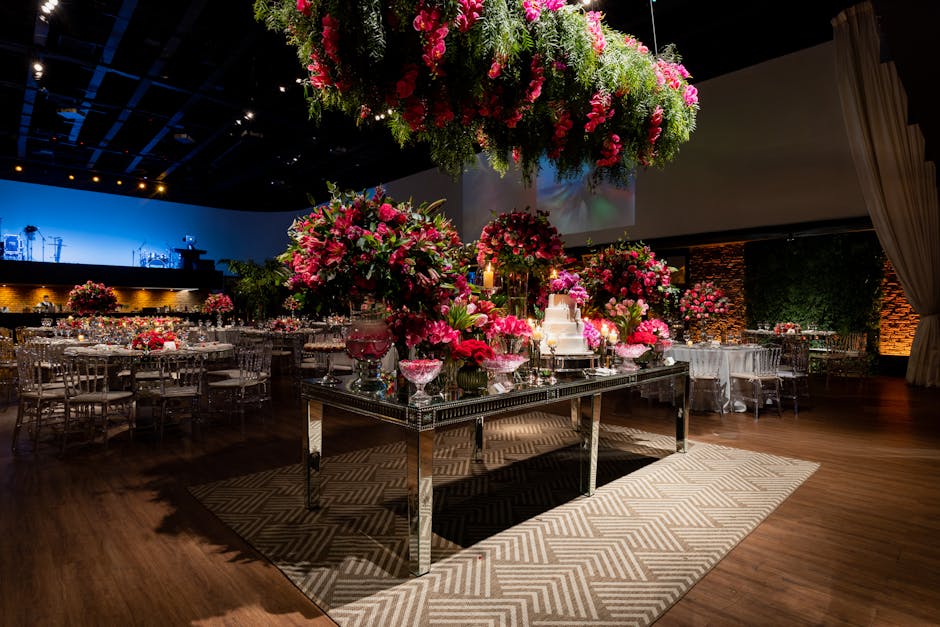 A floral arrangement display featuring large, vibrant bouquets predominantly composed of pink roses, peonies, and lilies, arranged in tall glass vases and complemented by lush greenery. The arrangemen