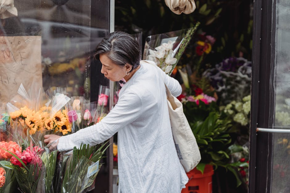 A woman with short, dark hair wearing a light grey coat and standing outside a florist shop, selecting a vibrant bouquet of fresh flowers wrapped in clear cellophane. The bouquet features large pink a