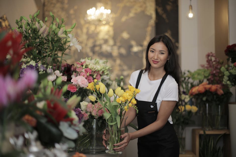A young woman with long dark hair, wearing a white t-shirt and black apron, is holding a tall, clear vase filled with fresh yellow tulips and green foliage. She is smiling gently and standing amid an 