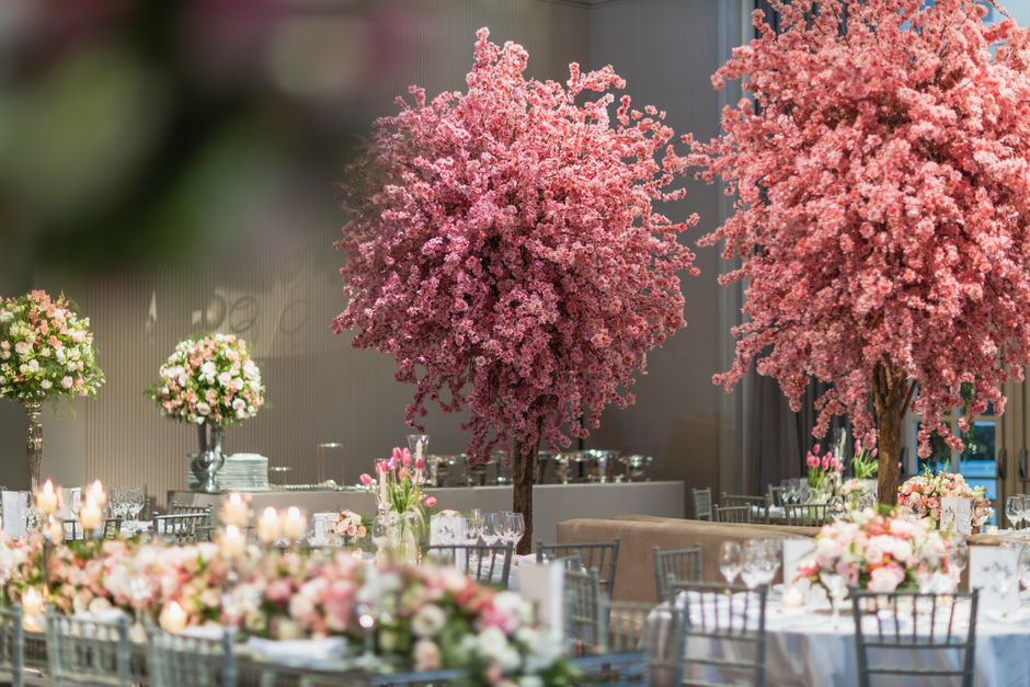 A sophisticated wedding reception setup featuring a floral arrangement with two large pink cherry blossom trees as the central focus, surrounded by elegant tables decorated with lush bouquets of pale 