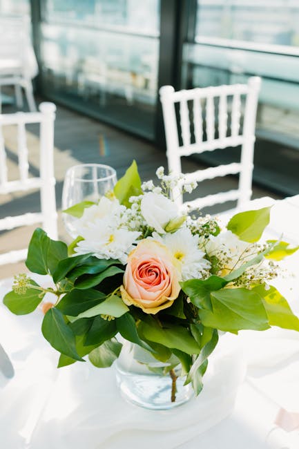 A floral arrangement in a clear glass vase featuring a large, soft pink rose at the center, surrounded by white peonies and small white filler flowers, accented with broad, glossy green leaves. The bo