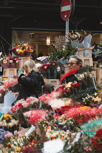 At a bustling flower market under a dark overpass, customers browse and select fresh floral arrangements from a variety of displays. The scene features vibrant bouquets of tulips, roses, lilies, and m