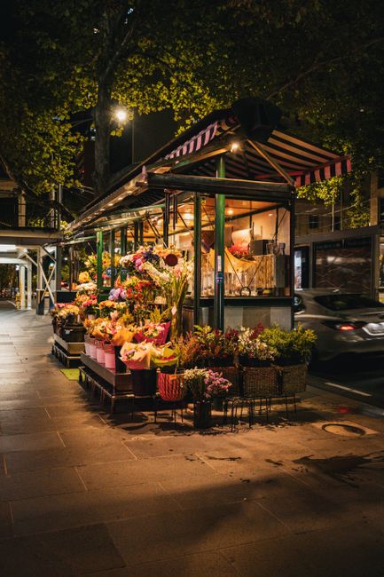 A flower stall located on a city street at night, illuminated by warm lighting from within the stall and a street lamp. The stall is covered by a red and white striped canopy and displays a vibrant ar